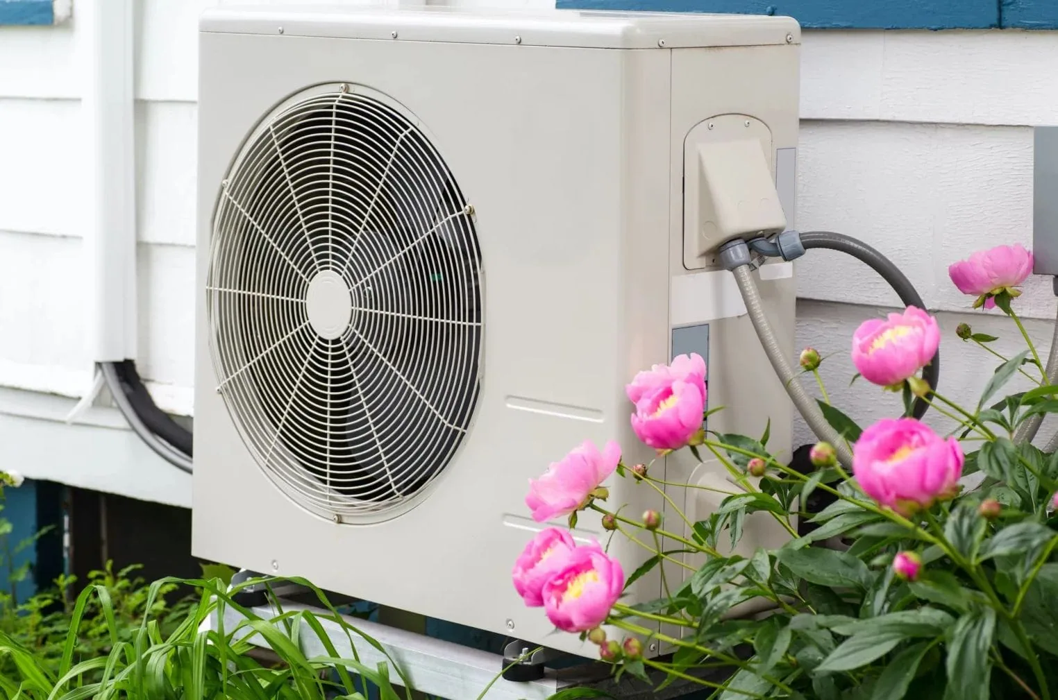 Outdoor heat pump unit beside a house with pink flowers in bloom